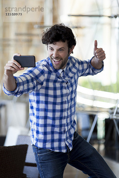 Ein Mann  der einen Daumen hochzieht und einen Selfie mit seinem Smartphone nimmt.