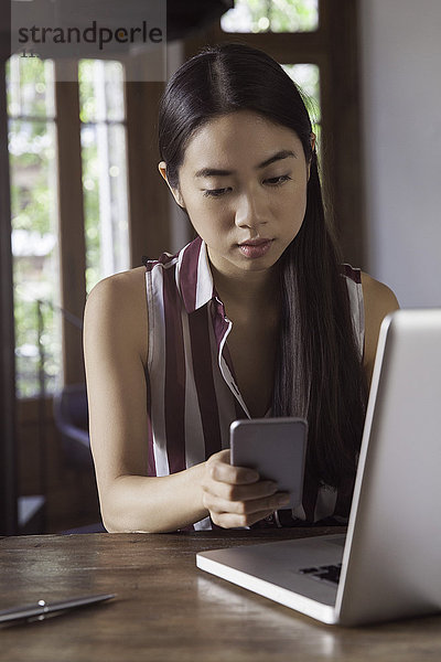 Junge Frau mit Smartphone zu SMS während der Arbeit am Laptop-Computer