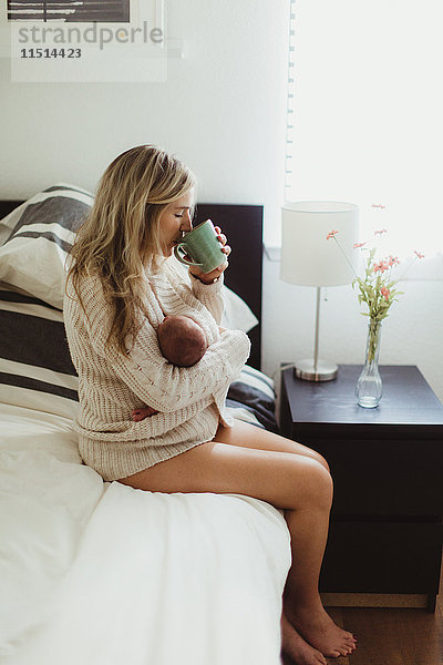 Mittlere erwachsene Frau sitzt im Bett und trinkt Kaffee  während sie ein neugeborenes Kind wiegt