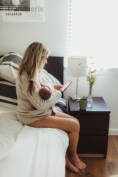 Mittlere erwachsene Frau sitzt auf dem Bett und schaut auf ein Smartphone  während sie eine neugeborene Tochter wiegt