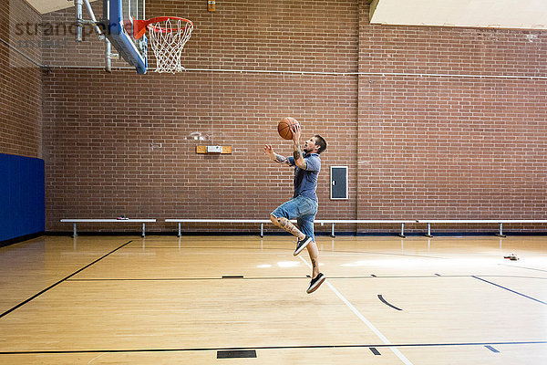 Tätowierter Mann springt und zielt mit dem Ball auf das Basketballnetz auf dem Platz