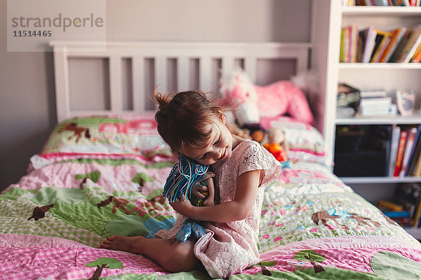 Mädchen sitzt auf Bett und umarmt Stoffpuppe