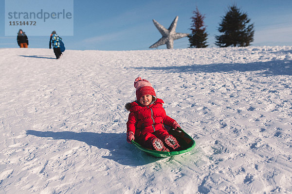 Mädchen mit roter Strickmütze rodelt einen schneebedeckten Hügel hinunter