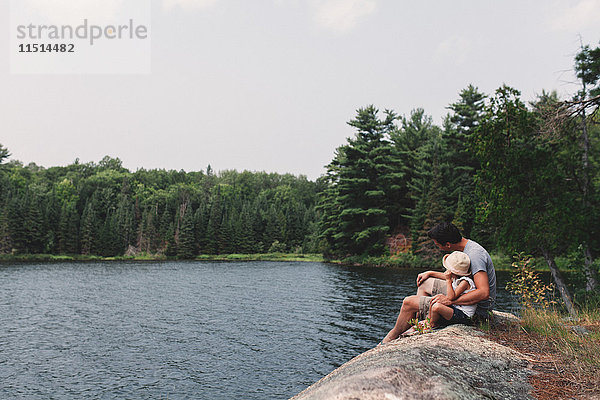 Junger Mann und Tochter sitzen auf einem Felsen am Rande eines Sees