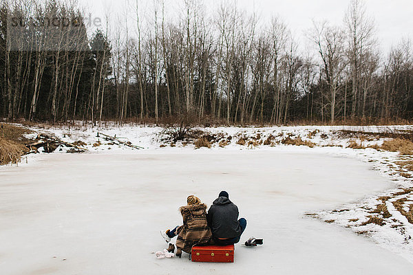 Paar sitzt auf rotem Koffer inmitten eines zugefrorenen Sees  Whitby  Ontario  Kanada
