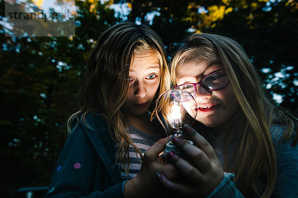 Zwei Mädchen  die in der Abenddämmerung im Garten eine beleuchtete Glühbirne halten und anstarren