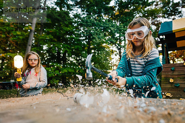 Mädchen zerschlägt Glühbirne mit Hammer auf Gartentisch in der Dämmerung