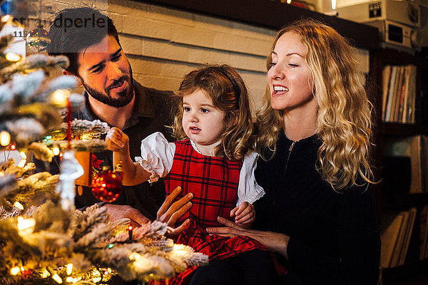 Weibliches Kleinkind mit Eltern  die eine Kugel auf den Weihnachtsbaum legen