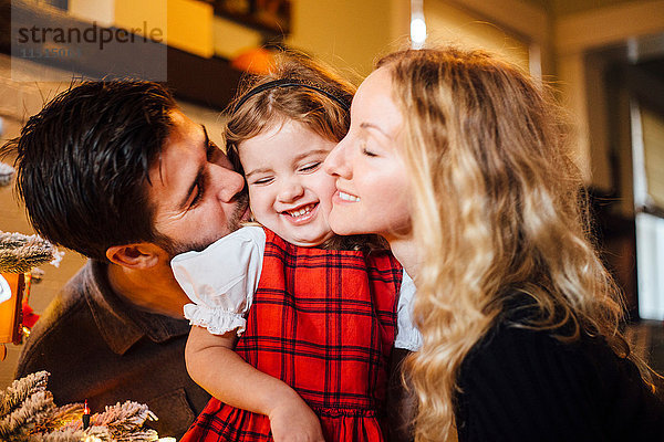 Weibliches Kleinkind zwischen Eltern  die es zu Weihnachten auf die Wange küssen