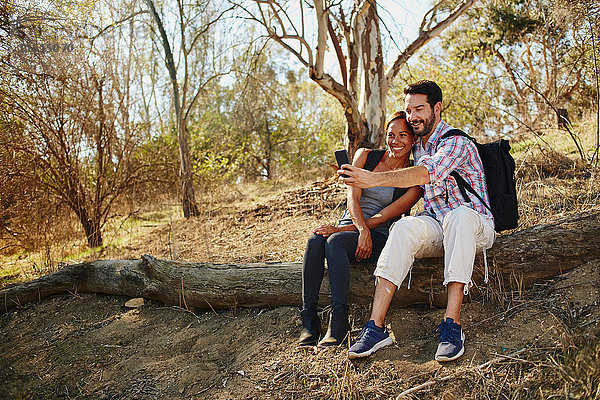 Mittelgroßes erwachsenes Paar beim Wandern  auf einem Baumstamm sitzend  Smartphone-Selfie