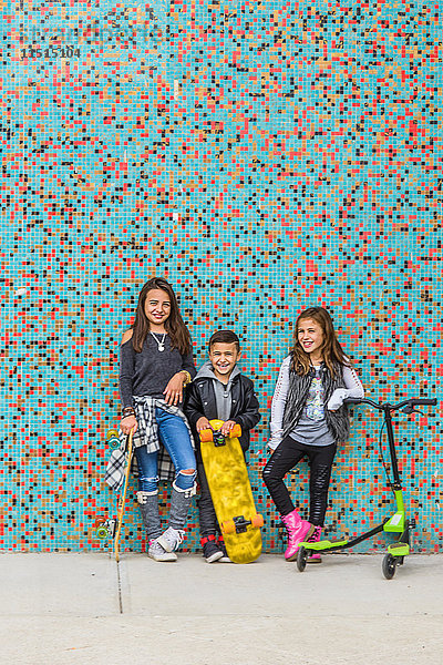 Porträt eines Jungen und zweier Schwestern mit Skateboards und Roller durch mehrfarbige Stadtmauer