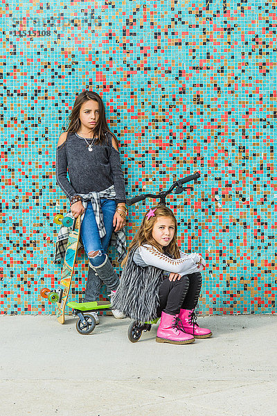 Porträt von zwei Schwestern mit Skateboards und Roller durch mehrfarbige Stadtmauer