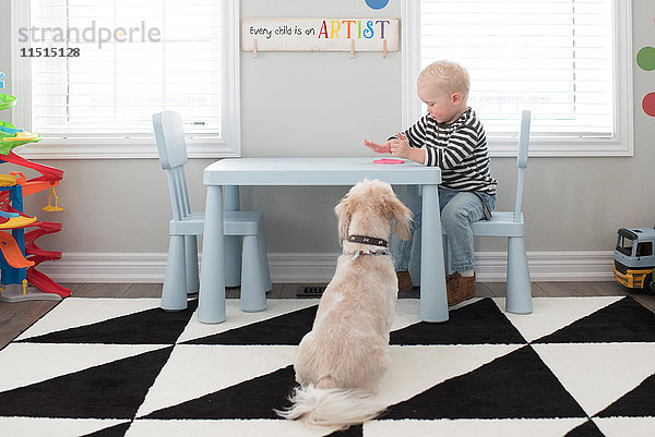 Junge spielt mit rosa Teig auf dem Tisch  Hund sitzt neben dem Tisch und schaut zu.