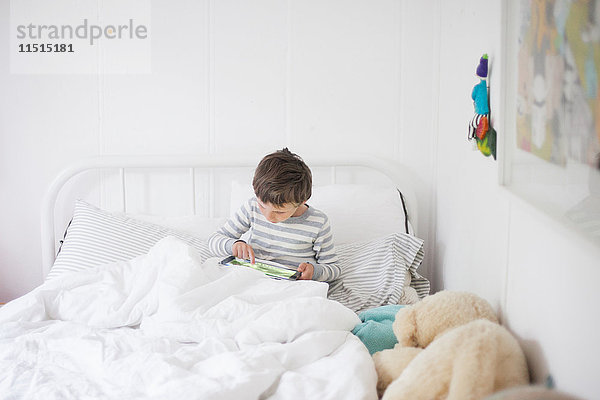 Aufrecht im Bett sitzender Junge spielt Videospiel auf digitalem Tablet