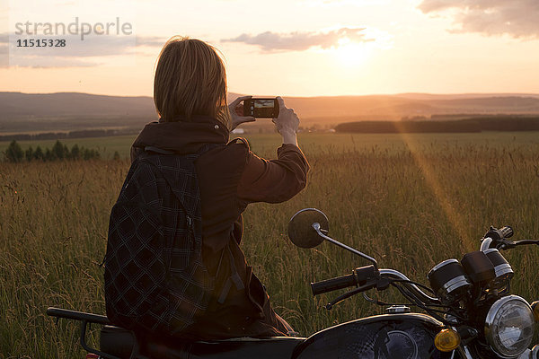Kaukasische Frau mit Motorrad im Feld beim Fotografieren des Sonnenuntergangs