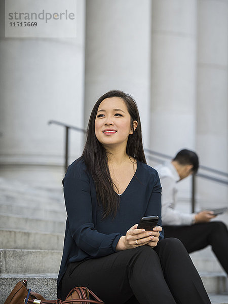 Chinesische Geschäftsfrau sitzt auf einer Treppe und hält ein Mobiltelefon in der Hand