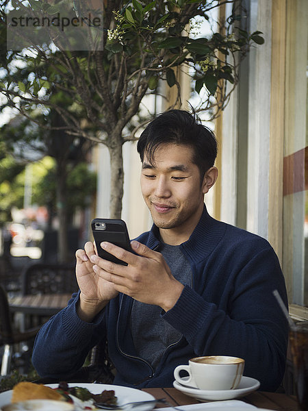 Lächelnder chinesischer Mann  der in einem Café im Freien eine SMS auf seinem Handy schreibt