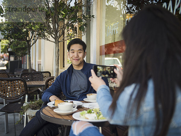 Chinesische Frau fotografiert Mann in einem Café mit Mobiltelefon