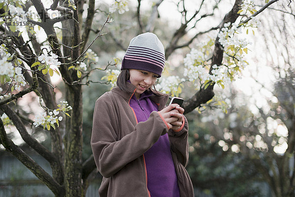 Lächelnde Mixed Race Mädchen texting auf Handy in der Nähe von Baum