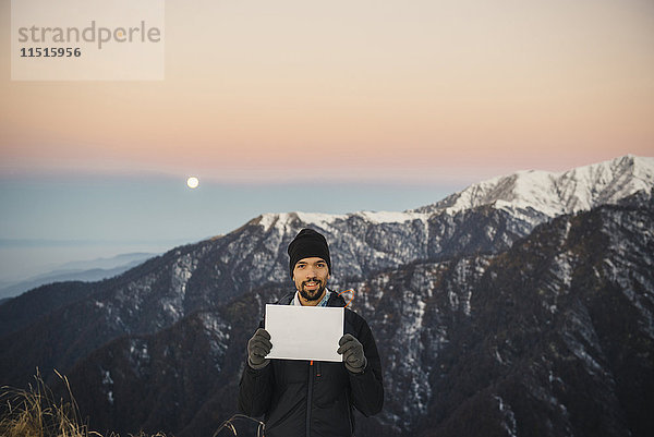 Lächelnder kaukasischer Mann hält leeres Schild in Berglandschaft