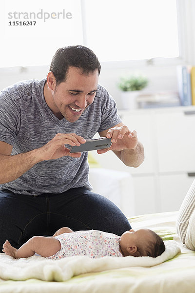 Hispanischer Vater auf dem Bett  der seine kleine Tochter mit dem Handy fotografiert