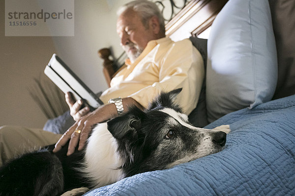 Älterer Mann liegt mit Hund auf dem Bett und liest auf einem digitalen Tablet
