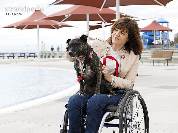 Frau im Rollstuhl sitzend am Wasser mit Hund auf dem Schoß