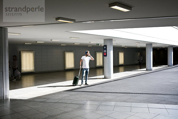 Junger Mann im Bahnhof  zieht Rollkoffer  trägt Kopfhörer  benutzt Smartphone