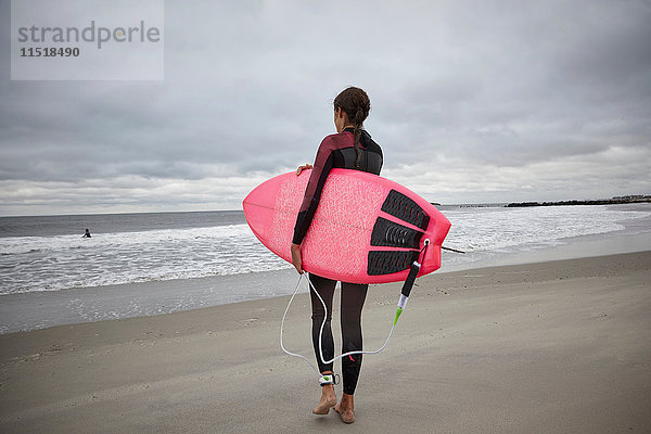 Rückansicht einer Surferin  die am Rockaway Beach  New York  USA  ein Surfbrett ins Meer trägt