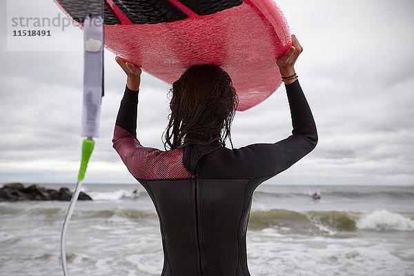 Rückansicht einer Surferin  die am Rockaway Beach  New York  USA  ein Surfbrett auf dem Kopf trägt