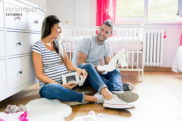 Schwangeres Paar sitzt auf dem Boden im Kinderzimmer und bereitet Babykleidung vor