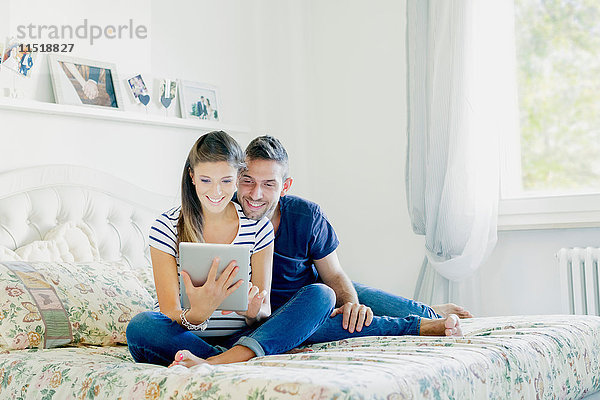 Ein schwangeres Paar sitzt auf dem Bett und benutzt ein digitales Tablet und lächelt