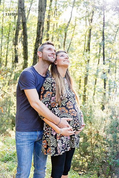 Schwangeres Paar im Wald umarmt sich  schaut lächelnd auf