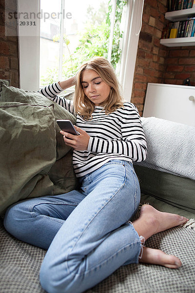 Junge Frau entspannt sich auf dem Sofa und schaut auf ein Smartphone