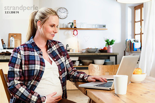 Schwangere Frau hält Bauch  während sie auf den Laptop auf dem Tisch schaut