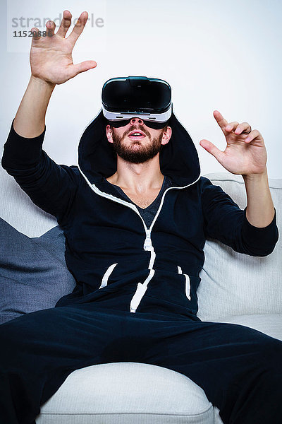 Mittelgroßer erwachsener Mann auf Sofa mit erhobenen Armen  der ein Virtual-Reality-Headset betrachtet