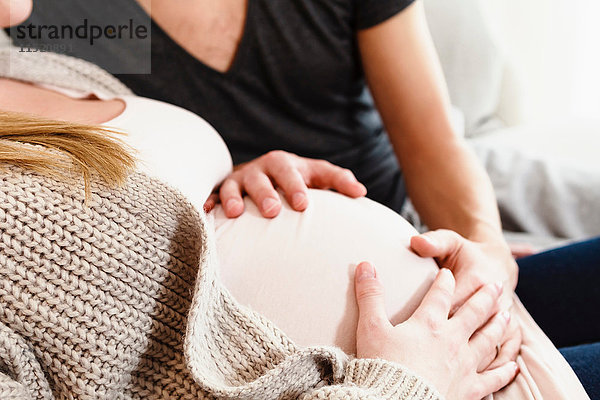 Schnappschuss eines schwangeren Paares im mittleren Erwachsenenalter  das den schwangeren Bauch auf dem Sofa berührt