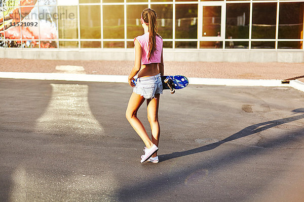 Junge Frau  die entlang der Straße geht  Skateboard trägt  Rückansicht