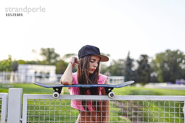 Junge Frau balanciert Skateboard auf Metallzaun  Ellbogen auf Skateboard