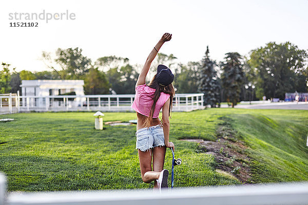 Junge Frau geht durch den Park  trägt Skateboard  schlägt Luft  Rückansicht