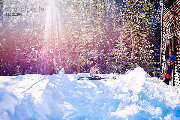 Frau praktiziert Yoga  kniend auf Yogamatte bei Blockhütte im Schnee  Österreich