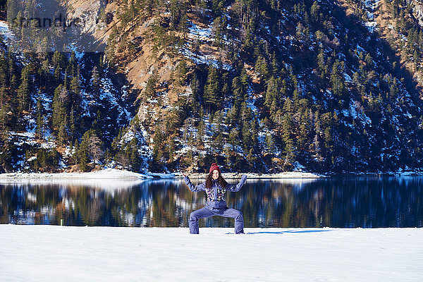 Frau in Winterkleidung praktiziert Yoga-Pose am verschneiten Seeufer  Österreich