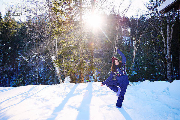 Frau in Winterkleidung praktiziert Baum-Yoga-Pose im Schnee am Wald  Österreich