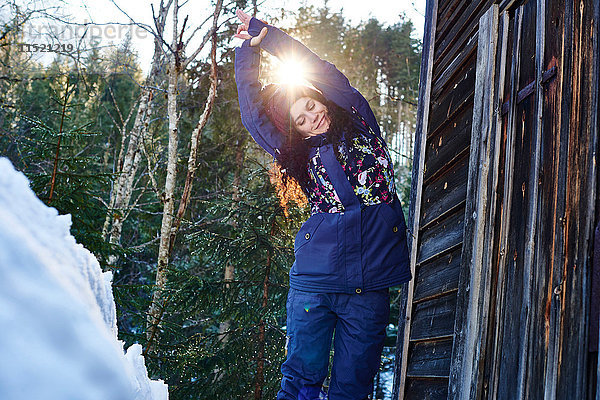 Frau in Winterkleidung übt Halbmond-Yoga-Pose im Schnee bei Blockhaus  Österreich