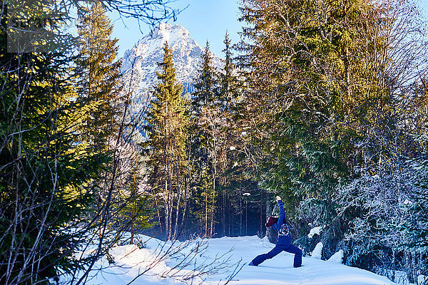 Frau in Winterkleidung praktiziert Krieger-Yoga-Pose im verschneiten Wald  Österreich