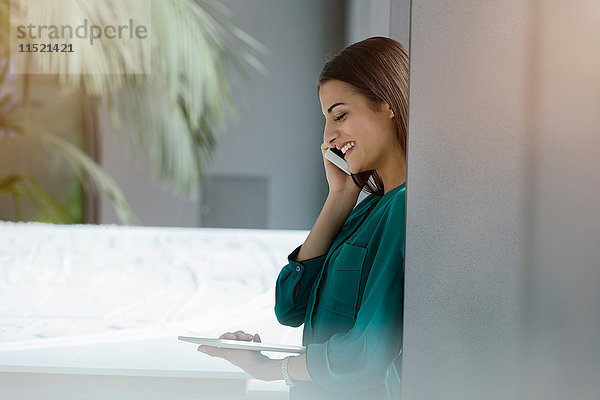 Junge Geschäftsfrau betrachtet digitales Tablet und spricht mit Smartphone im Büro