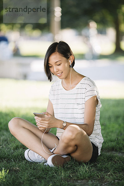 Lächelnde junge Frau sitzt auf einer Wiese und hört Musik mit Kopfhörern.