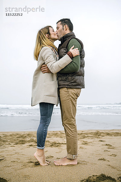 Ein Paar steht barfuß am Strand und küsst sich.
