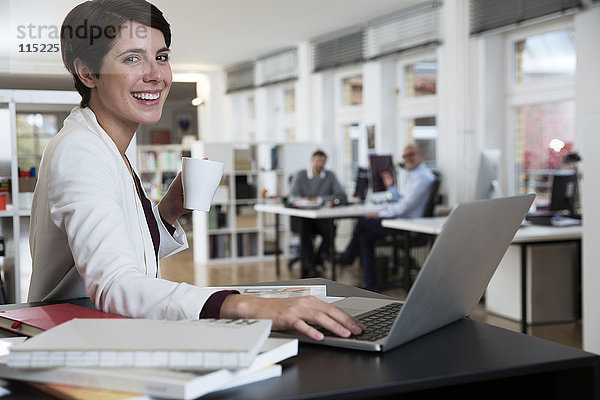 Porträt einer glücklichen Frau mit Laptop im Büro mit Kollegen im Hintergrund