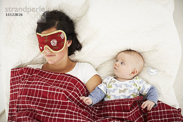 Mutter mit Schlafmaske und wachem Baby im Bett liegend